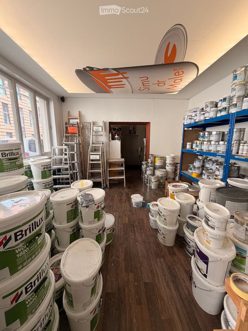 A well-lit room with wooden flooring, filled with various paint buckets and containers, shelves, and ladders.