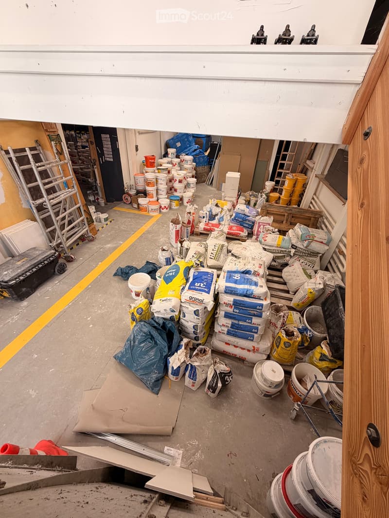 An unfurnished room filled with construction materials, including bags of cement, buckets, ladders, and tools, indicating a workspace for building or renovation.