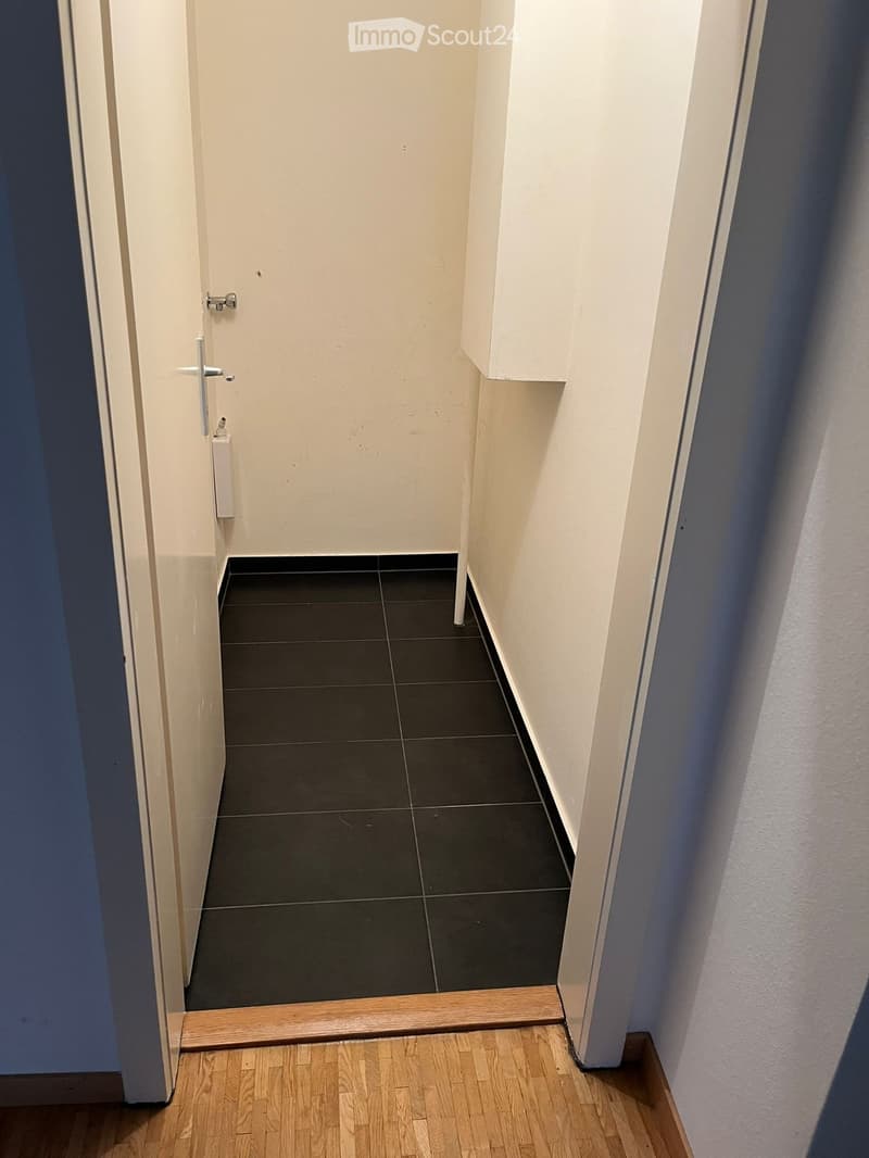 A narrow hallway with white walls, black tiled floor, and a wooden baseboard. The hallway leads to a door with a silver handle and a small white object attached to the wall beside it.