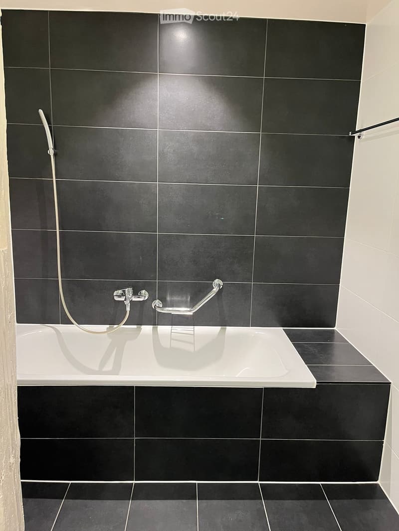 Modern bathroom with a white bathtub, black tiled walls and floor, a handheld shower hose, and a grab bar for safety.