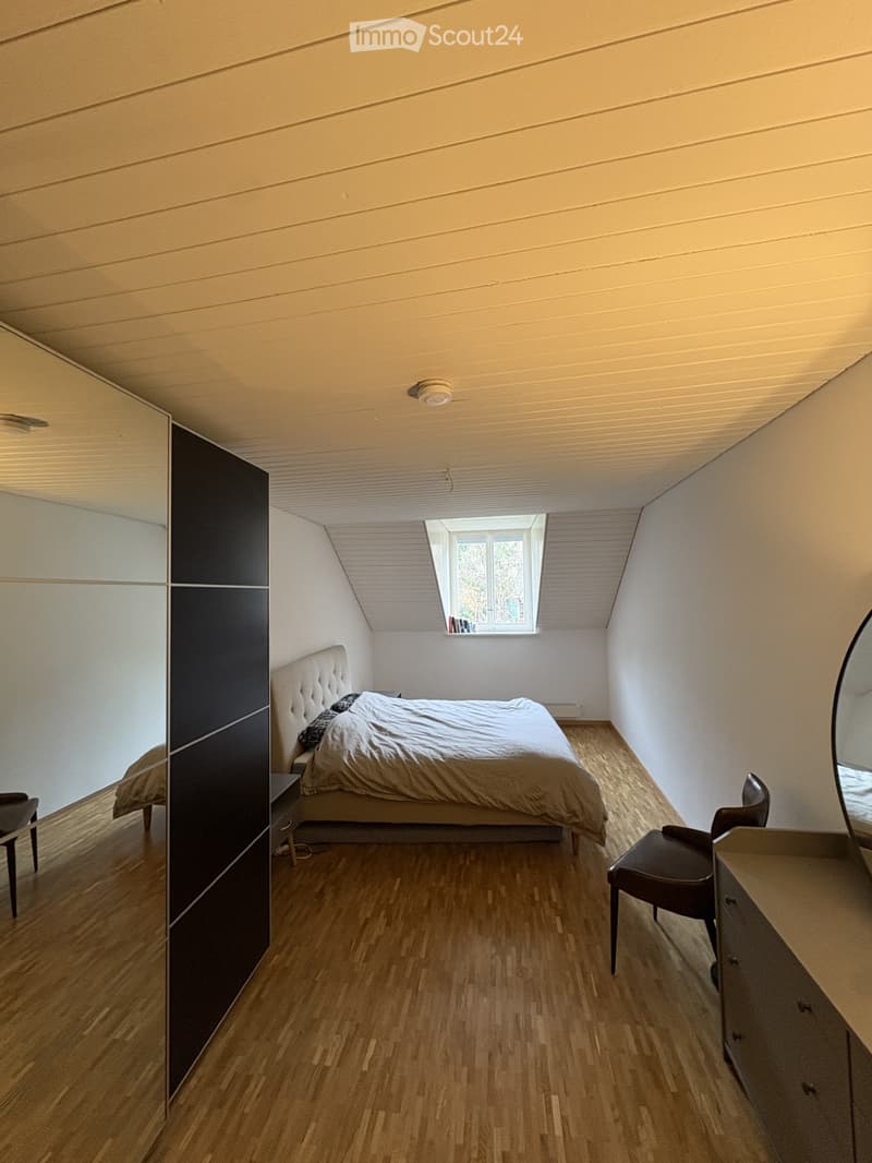 Modern bedroom with wooden flooring, a large bed with a tufted headboard, a mirrored wardrobe, a nightstand, and a small window with a view of greenery.