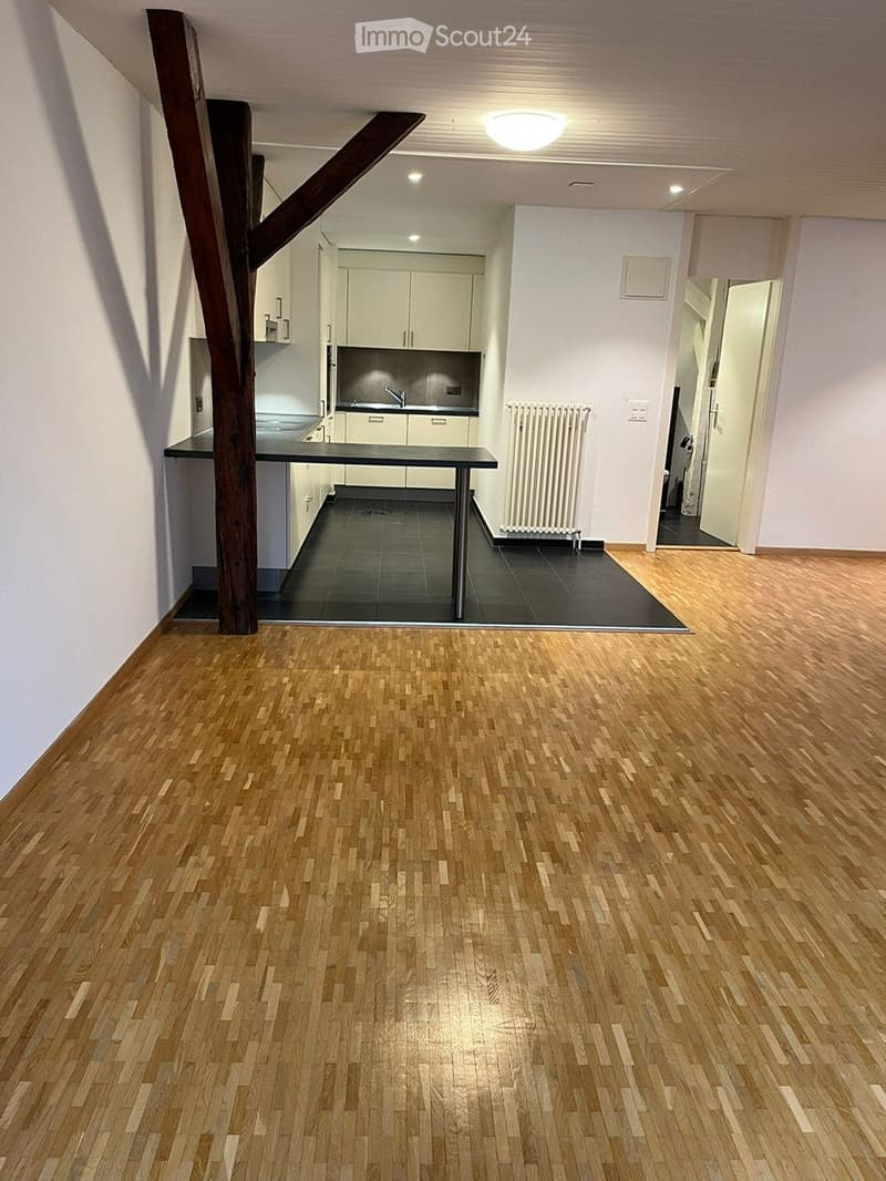 Empty room with wooden flooring, white walls, and a small kitchen area visible in the background. No furniture or furnishings are present.