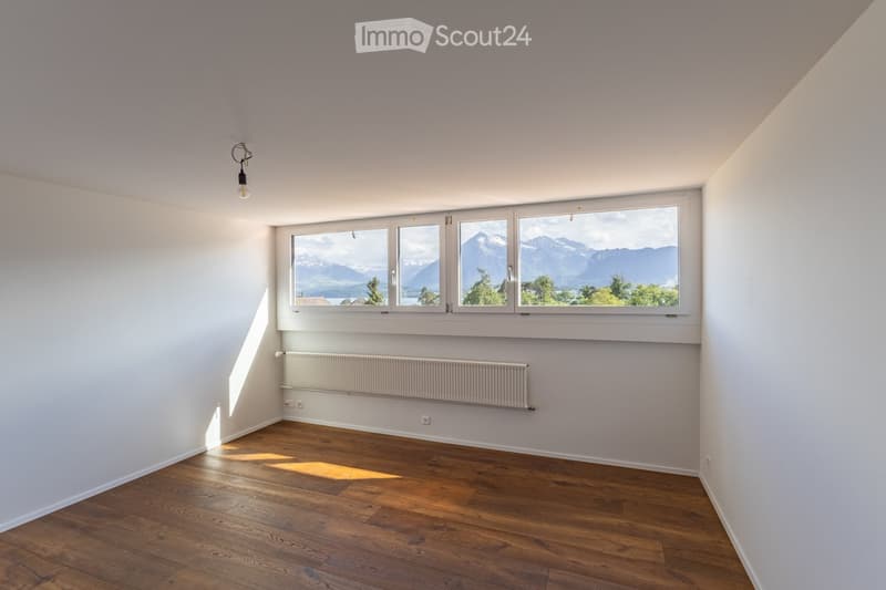 Empty room with wooden flooring, white walls, large windows offering a scenic mountain view, and a radiator below the windows.