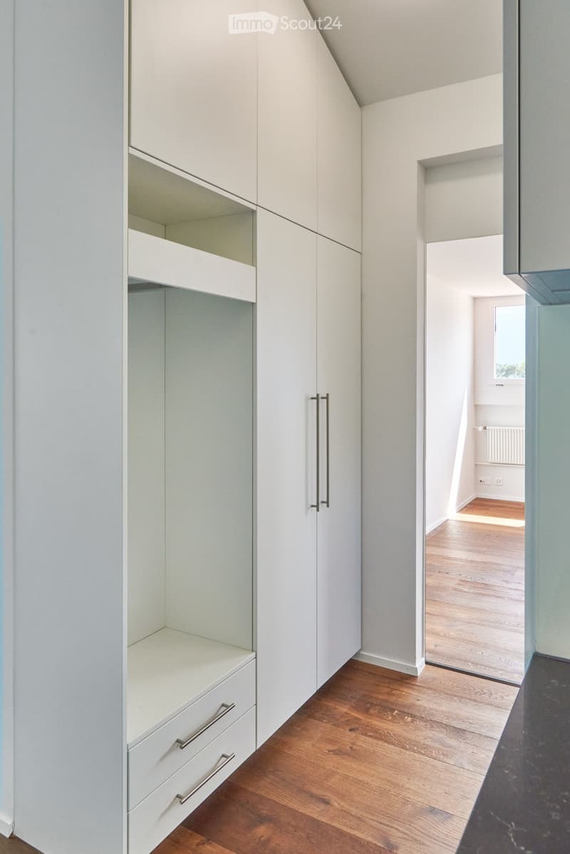 An empty room with white built-in cabinets and drawers, wooden flooring, and a view into another room with wooden flooring and white walls.