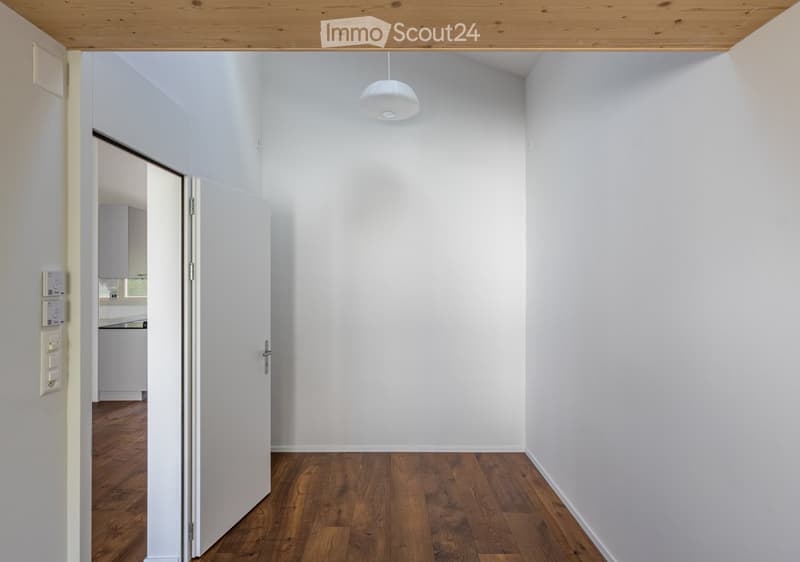 Empty hallway with white walls, wooden flooring, and a pendant light fixture. An open door leads to another room with cabinets and a countertop.
