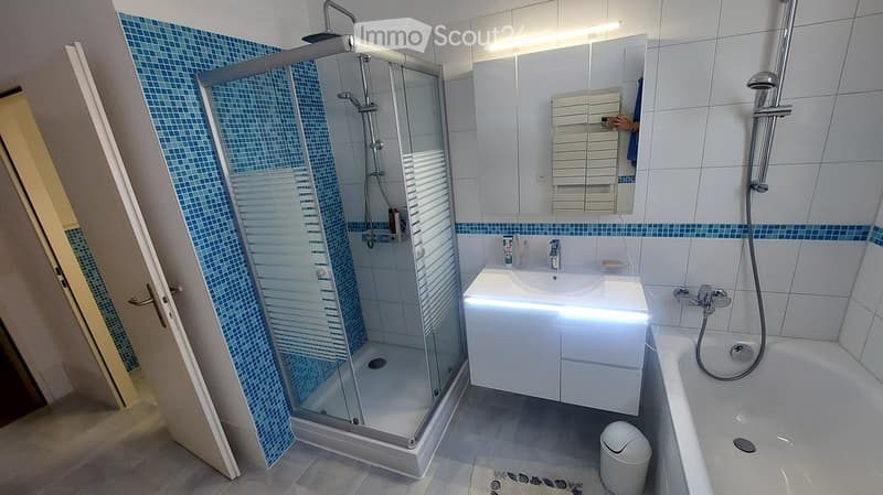 Modern bathroom with a glass-enclosed shower, white sink with built-in storage, bathtub, and a large mirror with built-in lighting. Blue mosaic tiles accent the shower and lower walls.
