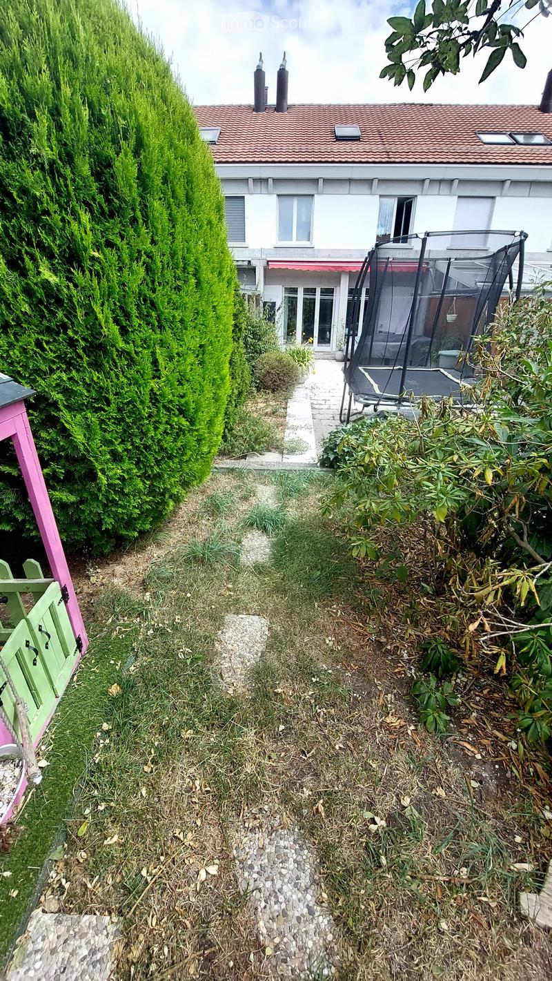 A narrow garden path leads to a house with a trampoline in the background. The garden features a tall, trimmed hedge on one side and various plants and shrubs on the other.