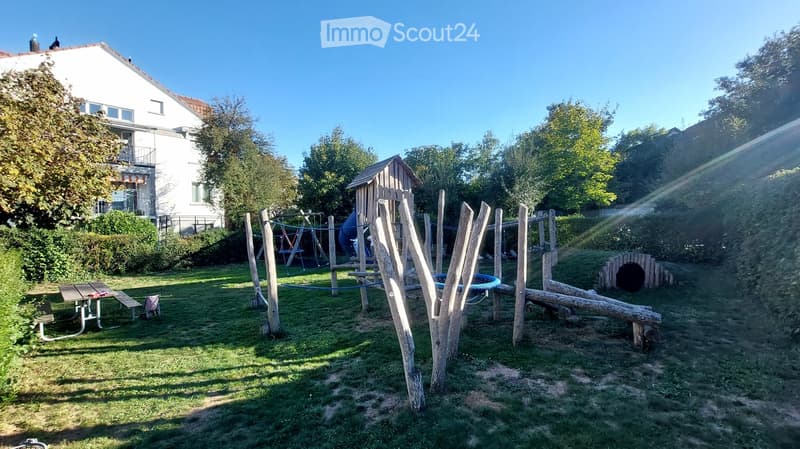A grassy backyard with a wooden play structure, a small wooden house, a trampoline, and a picnic table. The area is surrounded by trees and a white house with a balcony is visible in the background.