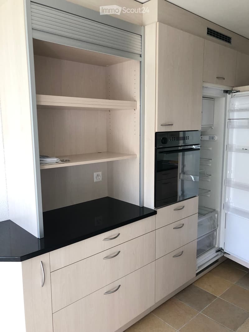 Modern kitchen with light wood cabinets, black countertop, open shelving, and stainless steel appliances including a refrigerator and oven.