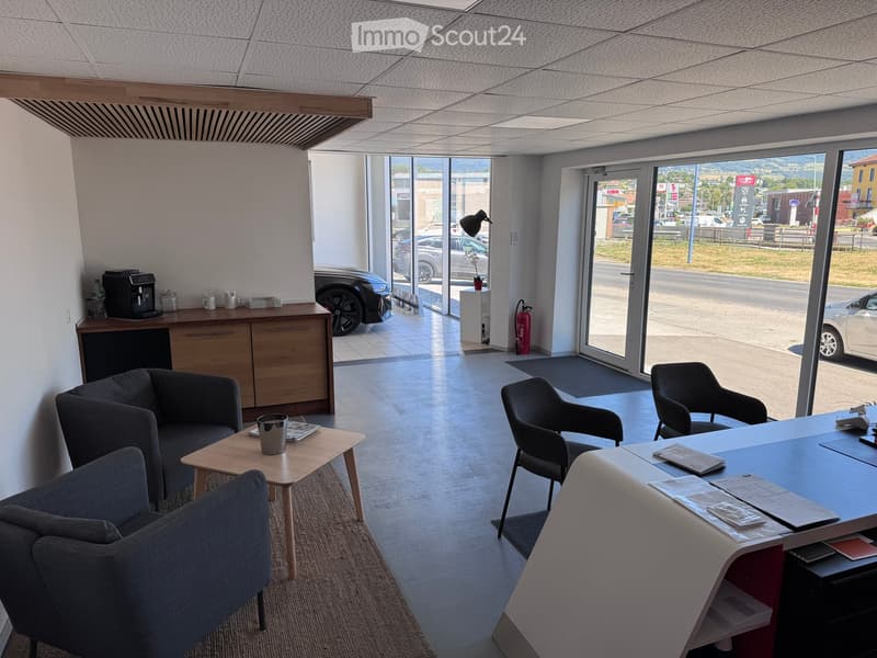 Modern office space with a reception area, featuring a sofa, chairs, a small coffee table, a desk, and a coffee machine. Large windows provide natural light and a view of the parking area outside.