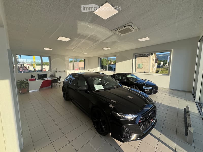 Car showroom with two black cars, a reception area with a table, chairs, a screen, and a potted plant.