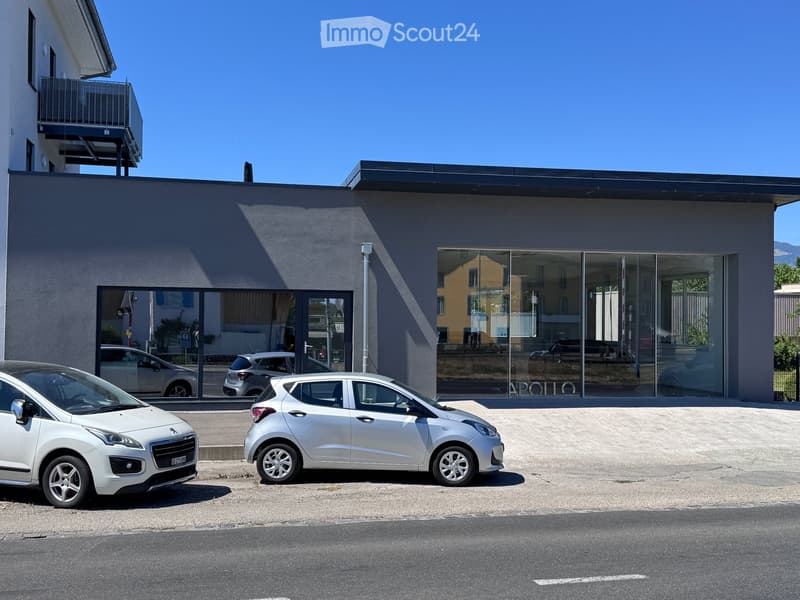 Modern two-story building with large glass windows and a flat roof, featuring a small balcony on the left side. Two cars are parked in front of the building.
