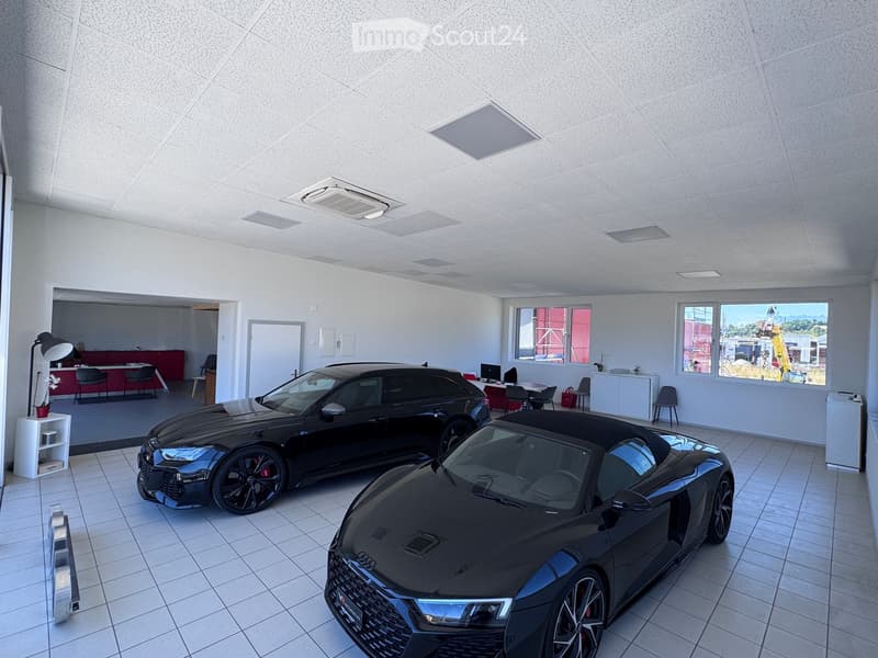 Modern showroom with two luxury cars, white walls, tiled floor, and a reception area with chairs, a table, and a lamp.