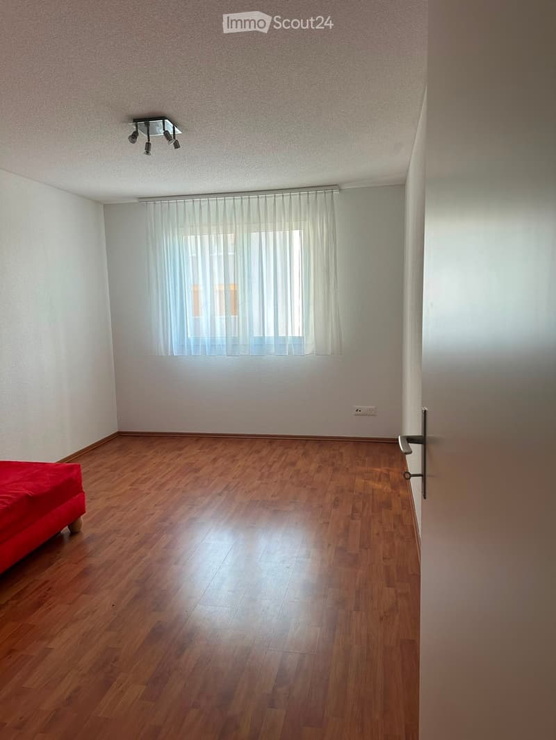Empty bedroom with wooden flooring, white walls, and a red bed visible on the left side.
