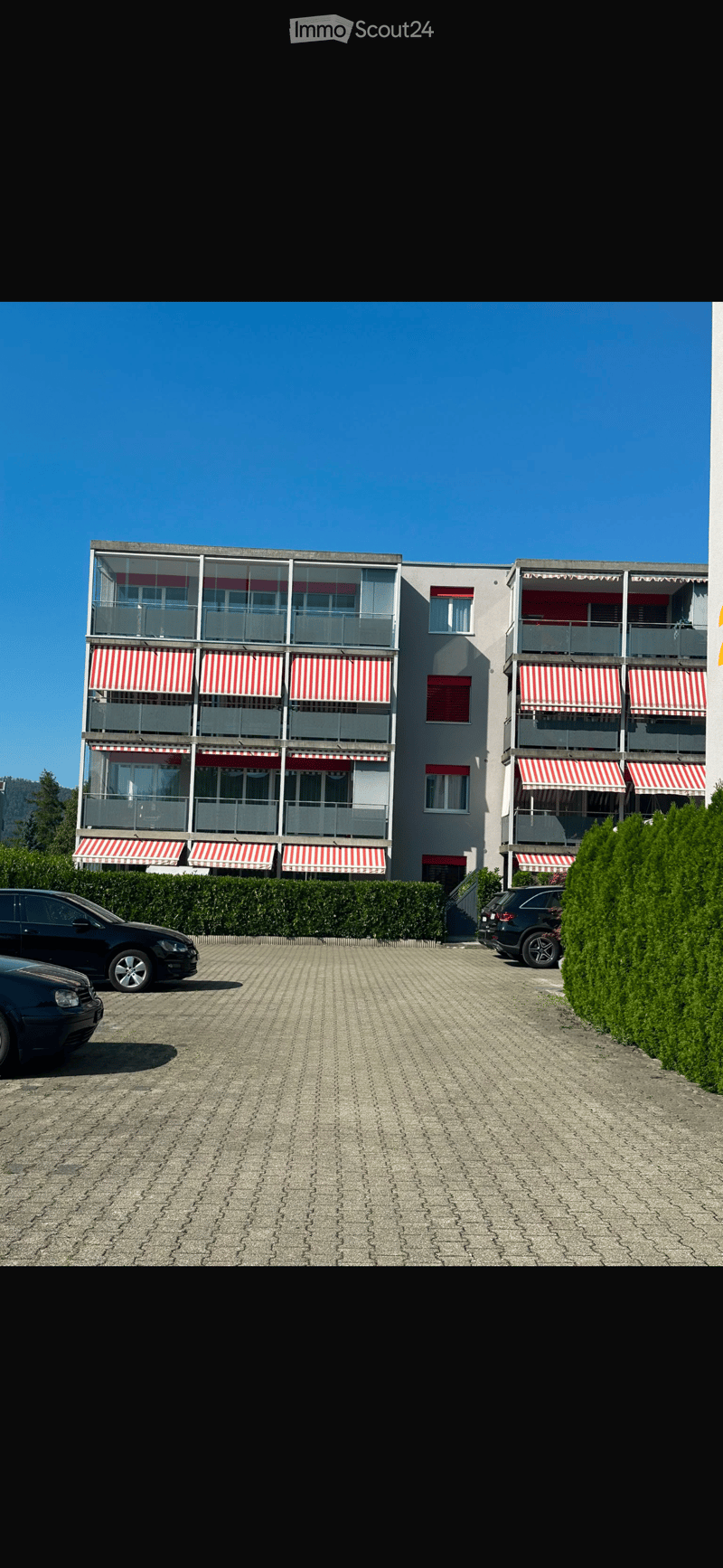 Modern multi-story building with balconies, striped awnings, and a paved parking area.