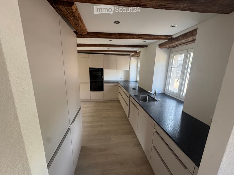 Modern kitchen with light-colored cabinets, dark countertops, and a stainless steel sink. Exposed wooden beams on the ceiling and large windows provide natural light.