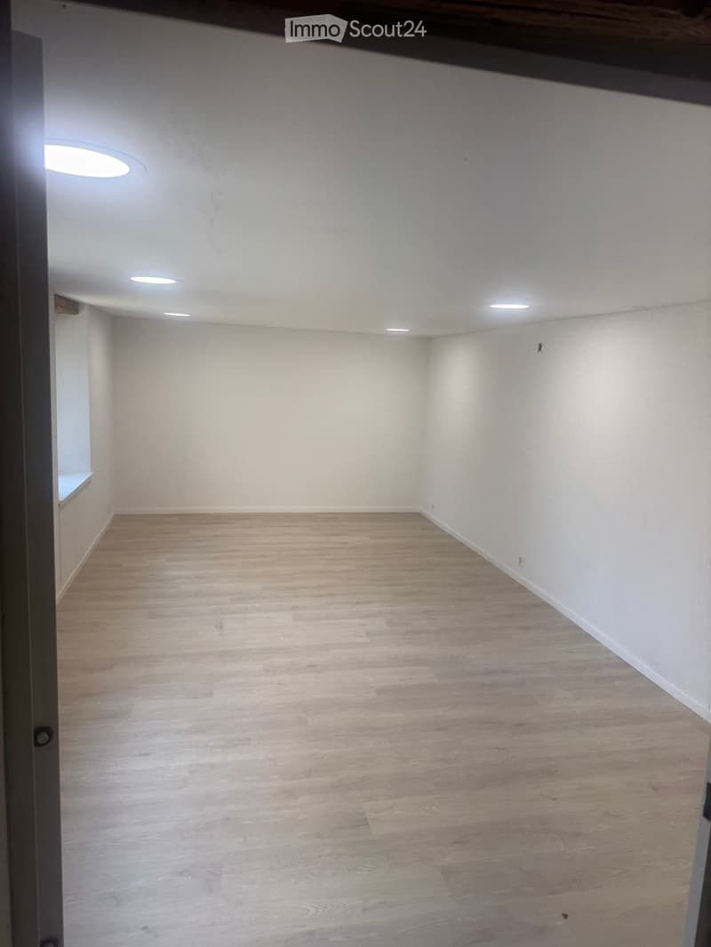 Empty room with light wooden flooring and white walls, featuring recessed ceiling lights and a small window on one side.