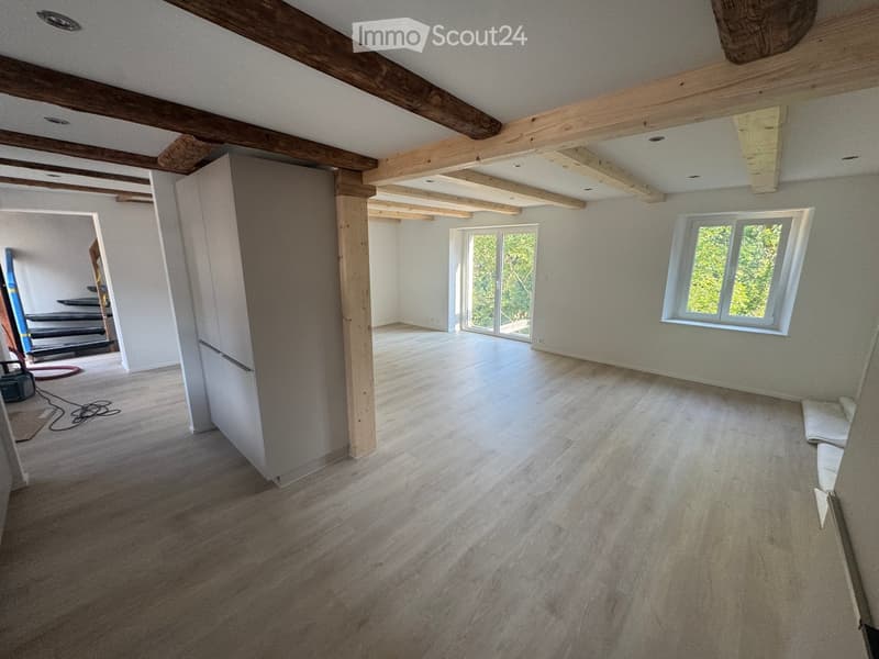 Empty room with exposed wooden beams, wooden flooring, and large windows letting in natural light. No furniture or equipment present.
