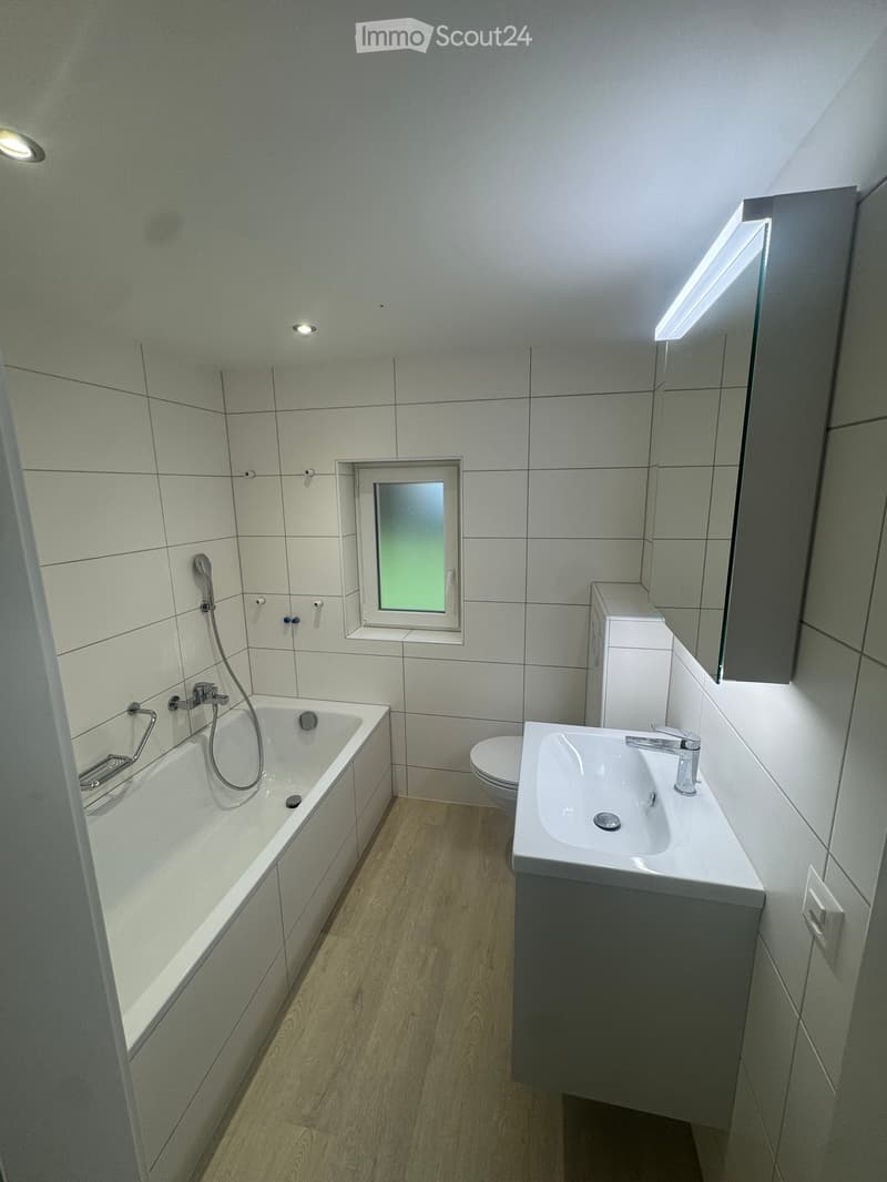 Modern bathroom with white tiled walls, a bathtub, a sink with a modern faucet, a toilet, and a large mirror with built-in lighting. The room features recessed ceiling lights and a small window with frosted glass.