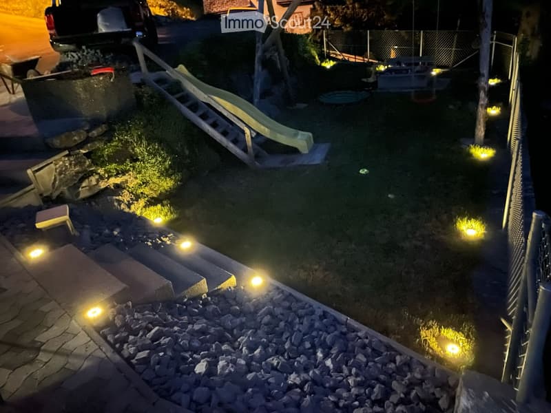 Nighttime view of a small garden area with a pool, surrounded by small lights, a lounge chair, and a fence.