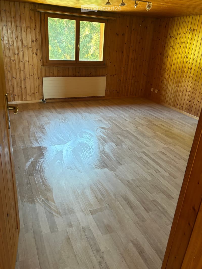 Empty room with wooden walls and floor, large window with wooden frame, radiator, and door with brass handle.