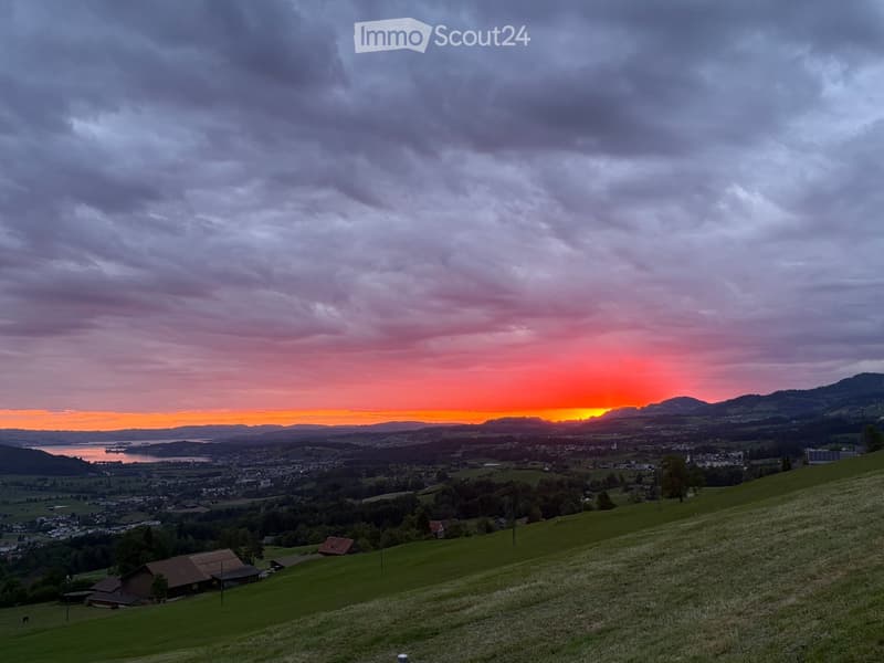 A panoramic view of a rural landscape at sunset, featuring rolling green hills, scattered houses, a body of water, and distant mountains under a dramatic sky.