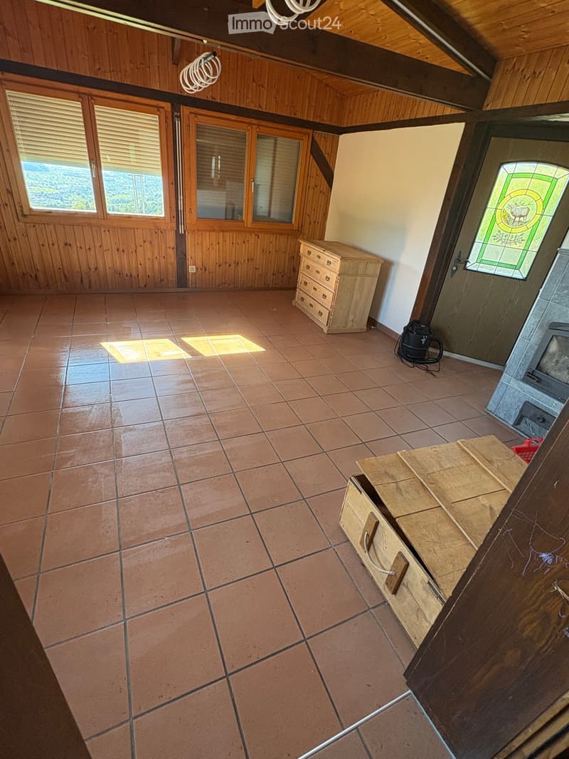 Empty room with tiled floor, wooden walls, and large windows. Features include a wooden chest of drawers, a small wooden bench, and a stained-glass window.