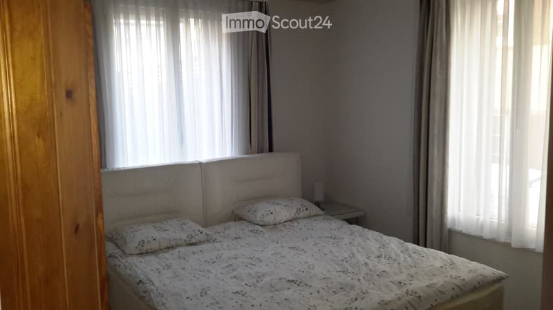 A neatly arranged bedroom with a double bed, patterned bedding, a headboard, and a nightstand with a lamp. The room has large windows with sheer curtains allowing natural light to fill the space.