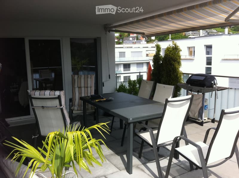 Balcony with a dining table, chairs, a grill, and a potted plant, featuring a retractable awning and a view of neighboring buildings.