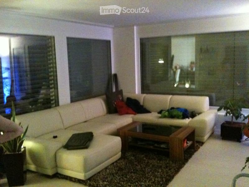 Living room with a white sectional sofa, coffee table, side table, and potted plants. Large windows with blinds are visible.