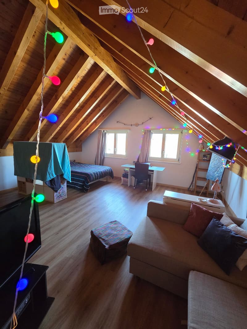 Cozy attic room with wooden beams, exposed ceiling, and colorful string lights. Features a sofa, ottoman, bed, desk, and television.