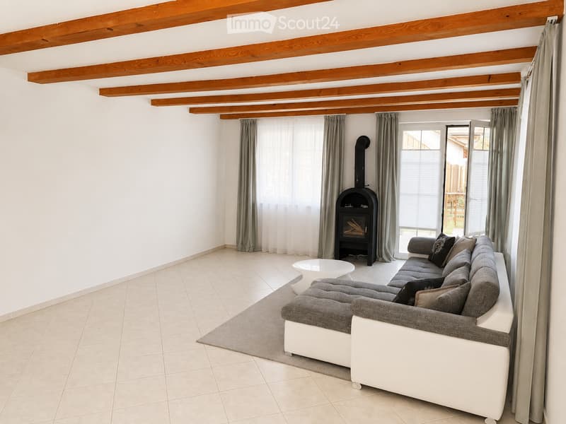 Bright living room with white walls, exposed wooden beams, large windows with grey curtains, grey sofa, white coffee table, and a black fireplace.