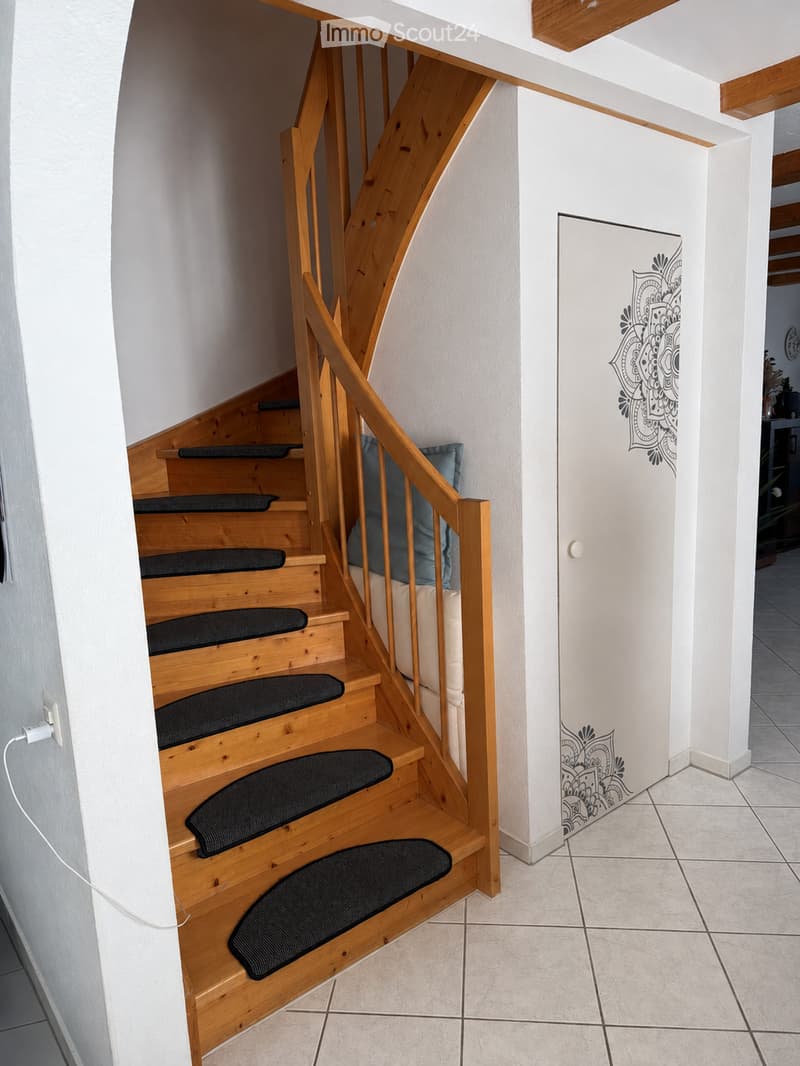 Wooden staircase with black carpeted steps and a wooden handrail, white walls with a decorative design, tiled floor, and a visible electrical outlet on the left side.