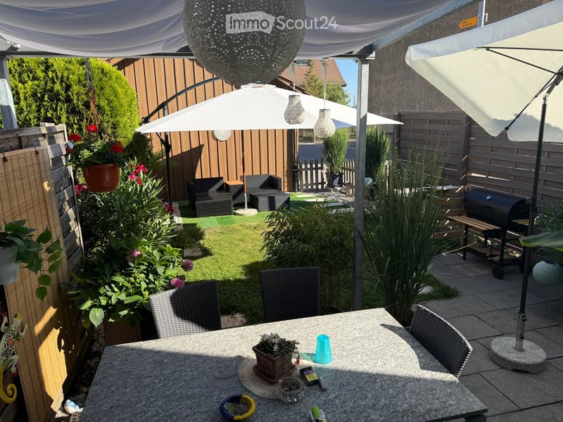 A well-maintained garden with a patio area featuring a stone table, chairs, umbrellas, potted plants, and a barbecue in the background.