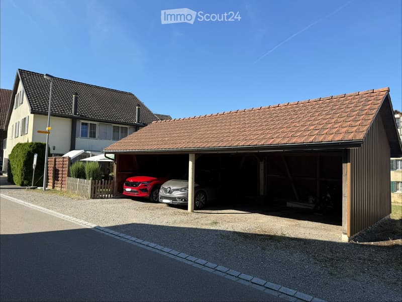 Two-story house with a tiled roof, a garage with two parked cars, and a small garden area with a fence.