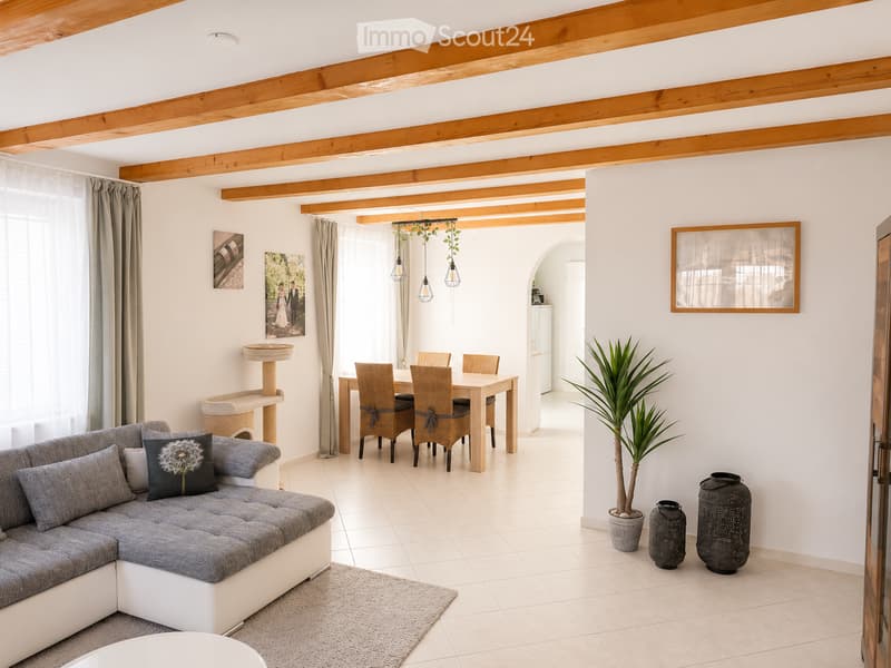 Bright living and dining area with exposed wooden beams, white walls, grey sofa, dining table with four chairs, cat tree, potted plant, and decorative vases.