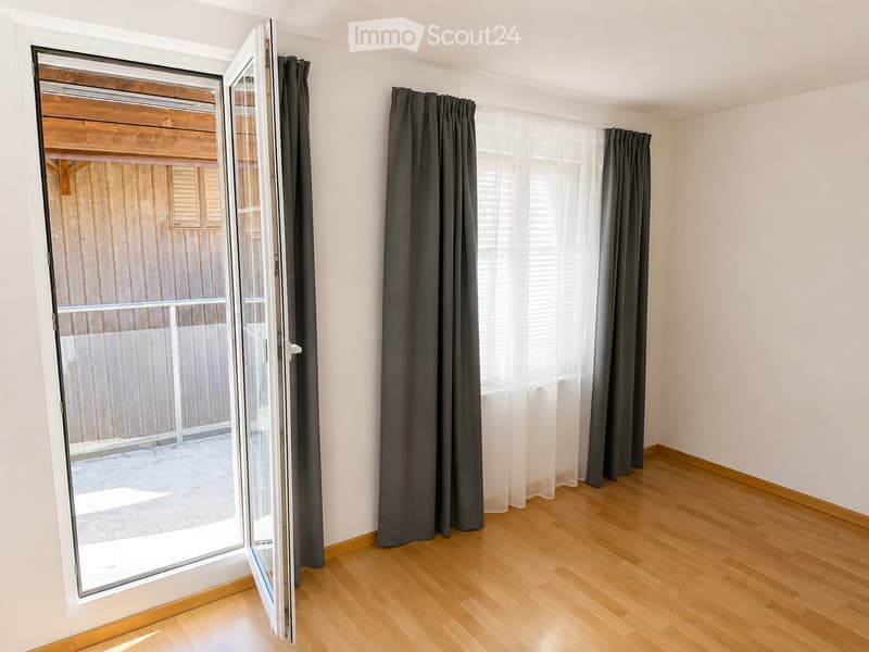 Empty room with wooden flooring, large window with dark curtains, and a sliding glass door leading to a wooden deck area.