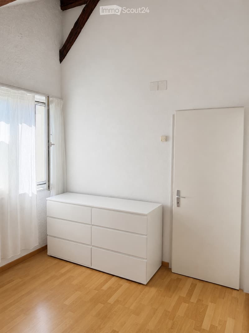 A small, unfurnished room with light wooden flooring, white walls, and a white dresser with multiple drawers. A white door is visible on the right, and a window with white curtains is on the left.