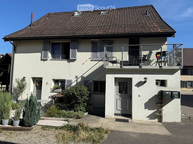 Two-story house with beige exterior, tiled roof, multiple windows with shutters, balcony with outdoor furniture, small garden area with potted plants and shrubs.