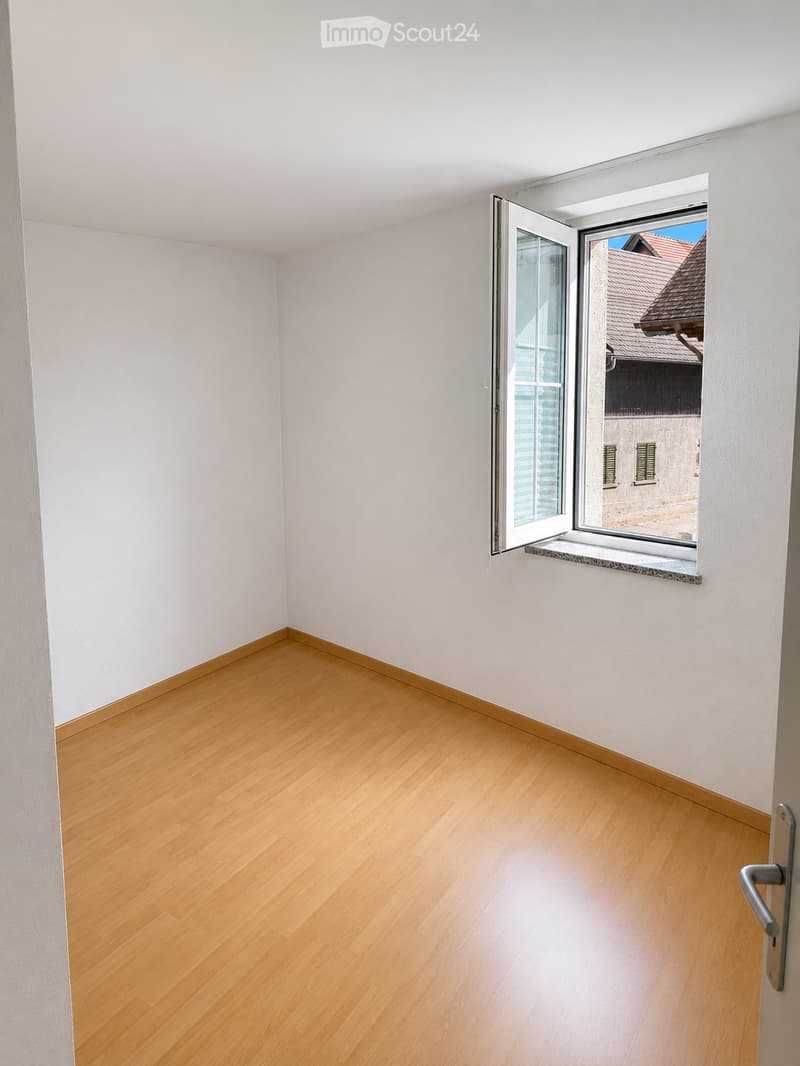 Empty room with light wooden flooring and white walls, featuring a large open window with a view of a neighboring building.
