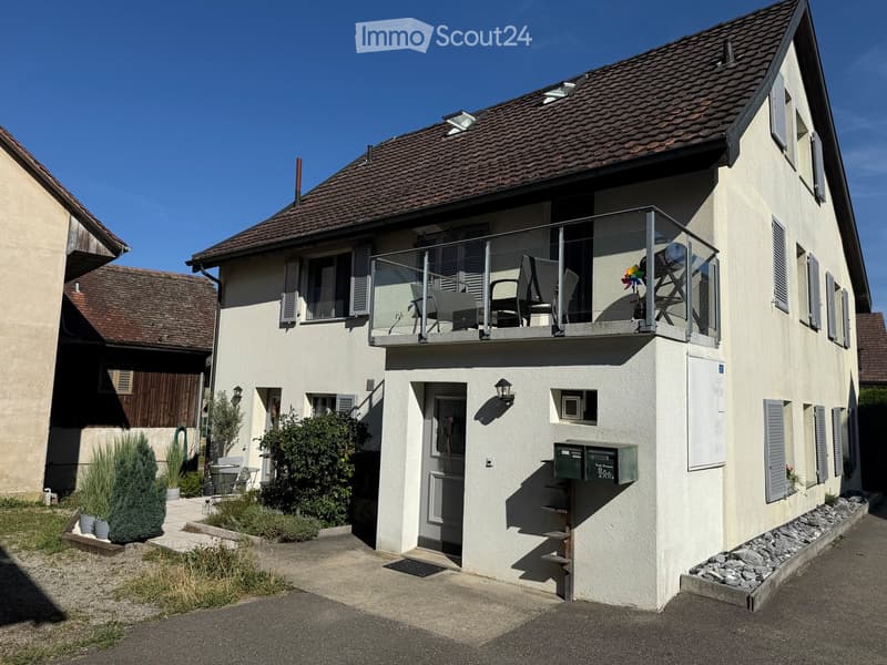 Two-story house with beige exterior, tiled roof, balcony with chairs, small garden, and mailbox.