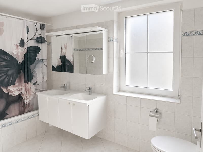 Modern bathroom with a double sink vanity, large mirror, toilet, and a shower with a decorative curtain. The room features tiled walls and a window for natural light.