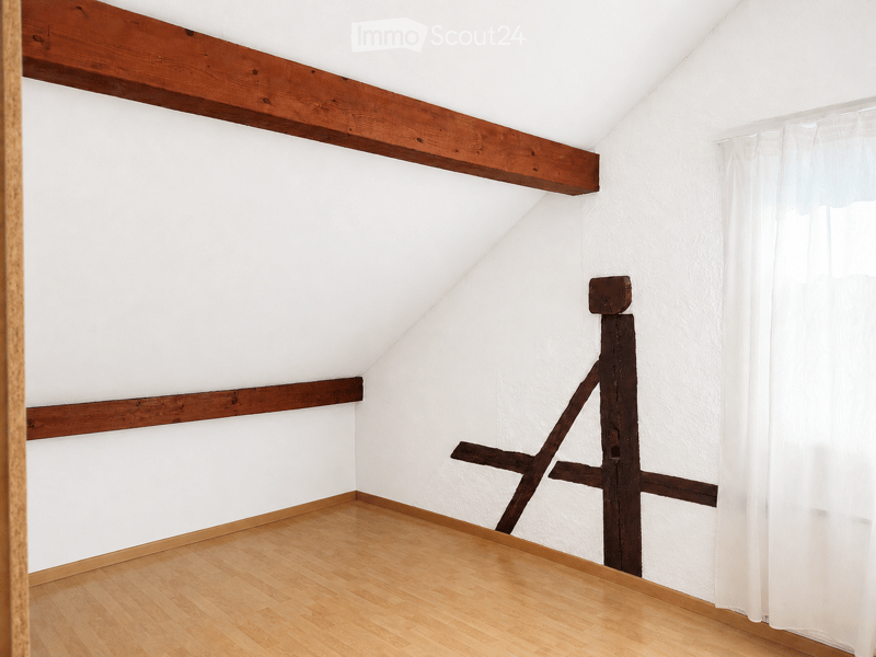 An empty attic room with exposed wooden beams, white walls, and a wooden floor. The room features a slanted ceiling and a window with white curtains.