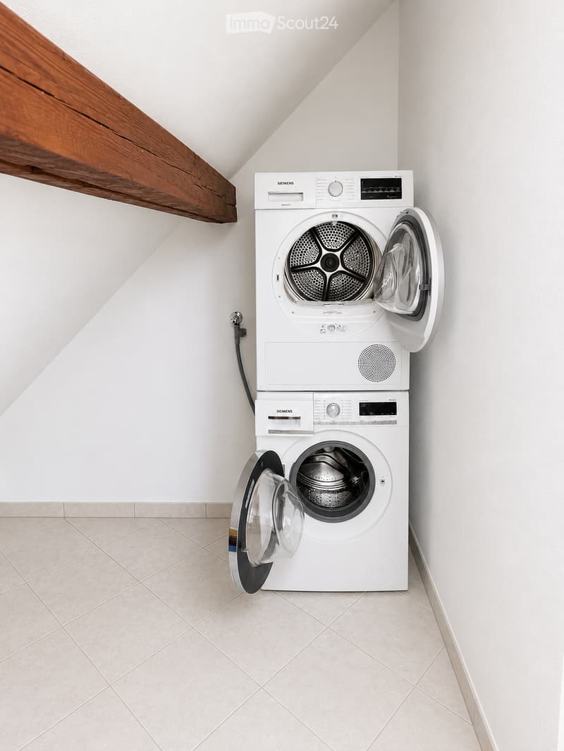 A modern bathroom with a white Siemens washing machine and dryer unit, both with open doors, placed against a white wall with a wooden beam above.
