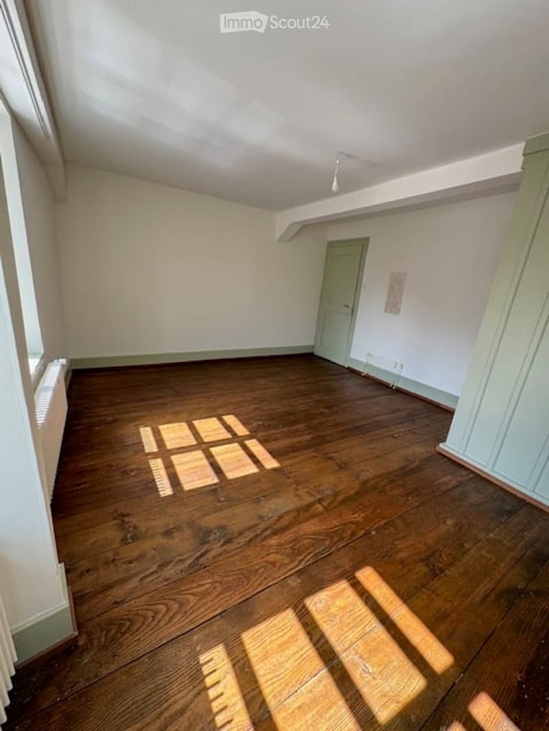 Empty room with wooden flooring, white walls, and a single door. Sunlight streams in from the left, casting shadows on the floor.
