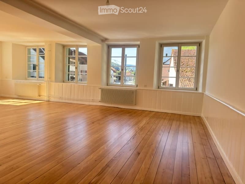 Empty room with wooden flooring, white walls, and multiple windows allowing natural light. Radiators are visible under the windows.