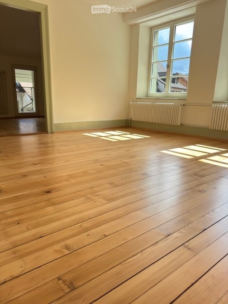Empty room with wooden flooring, large window with natural light, radiator, and visible view of another room with a balcony.