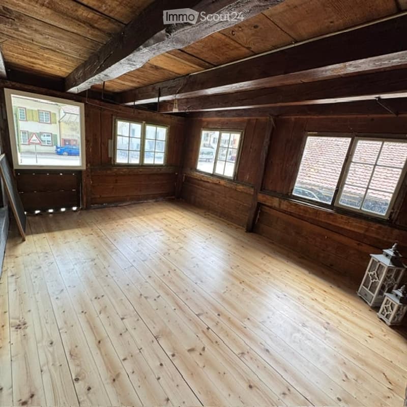 Empty wooden room with exposed wooden beams on the ceiling, large windows, and a view of the street outside. The floor is made of wide wooden planks.