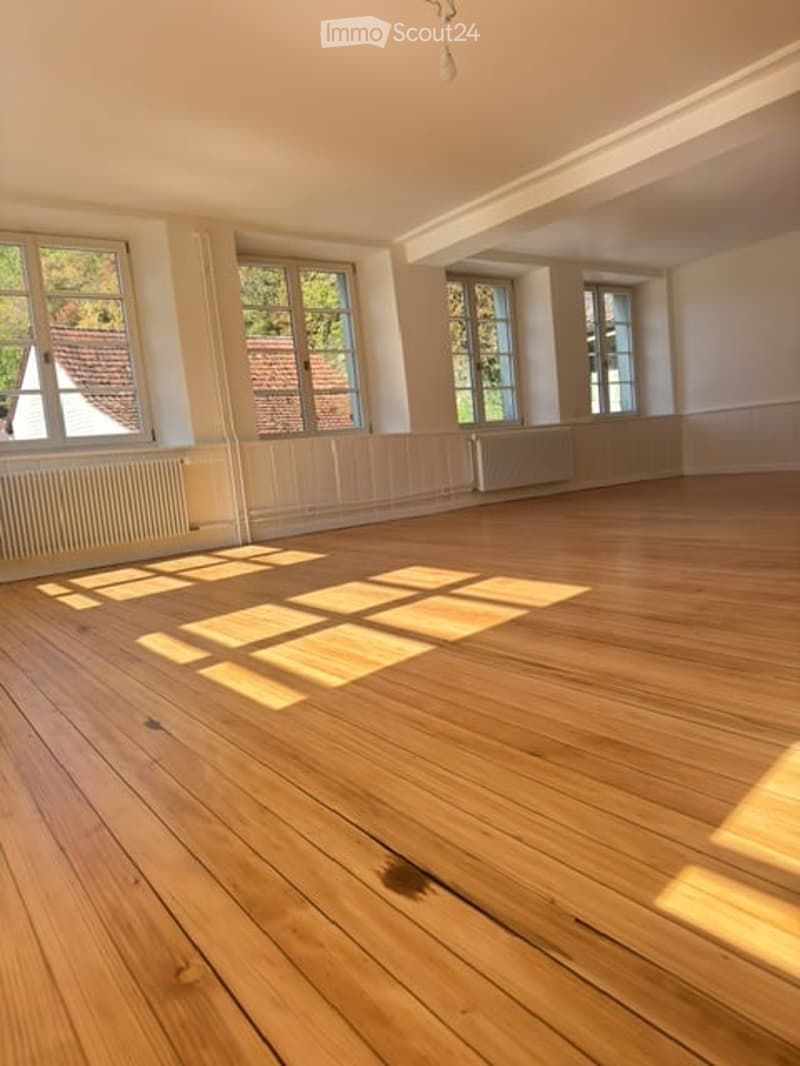Empty room with wooden flooring, white walls, and large windows allowing natural light to enter. No furniture or equipment is present.