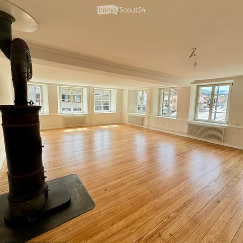 Empty room with wooden flooring, large windows, and a central heating stove. No furniture or equipment present.